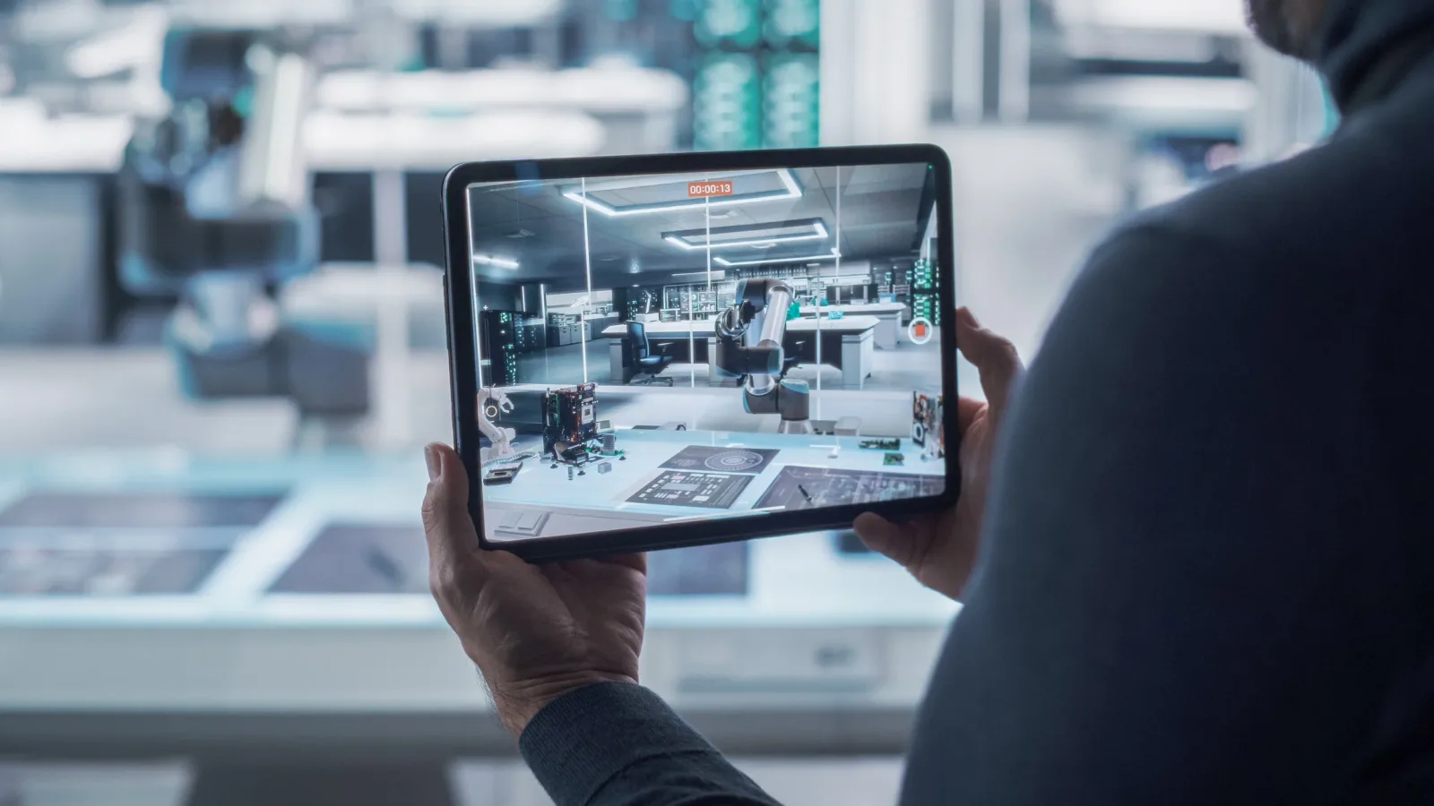Person filming a robotics laboratory with a tablet, showing robotic arms and electronic components on a worktable.